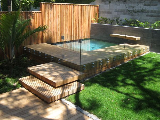 A landscaped backyard with a compact swimming pool surrounded by a raised timber deck and transparent glass balustrades. A timber privacy fence, dark rendered masonry wall, built-in bench, stepped timber platforms, tropical planting and lawn create a contemporary outdoor entertaining area.