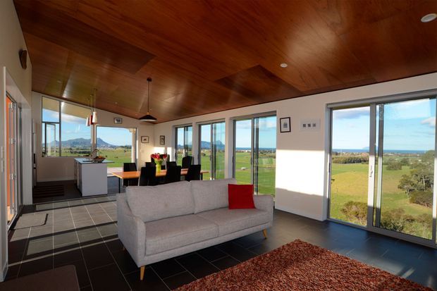 Bright open-plan living and dining space with a compact kitchen, expansive floor-to-ceiling glazing, dark tiled flooring, a warm timber-lined ceiling, and views across landscaped grounds toward the ocean. A light grey sofa and red accent cushion sit on a textured rust-coloured rug, while a dining table and kitchen island occupy the rear of the room.