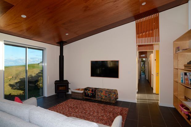 A warm contemporary living room with a rich timber ceiling, white walls, dark tiled flooring, and a freestanding black wood-burning stove. The room includes a large sofa, patterned ottoman, textured rug, wall-mounted television, built-in shelving, and a tall slatted timber screen opening to a hallway. Full-height glazing provides expansive coastal landscape views.