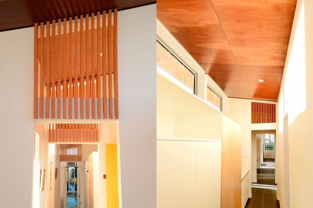 Bright contemporary interior hallway with warm timber ceilings and repeated vertical timber slat screens forming a decorative balustrade or partition. White walls and built-in cabinetry contrast with dark tiled flooring, while the long corridor leads toward glazed doors and additional rooms. Strong natural light and clerestory glazing create an airy, linear space.