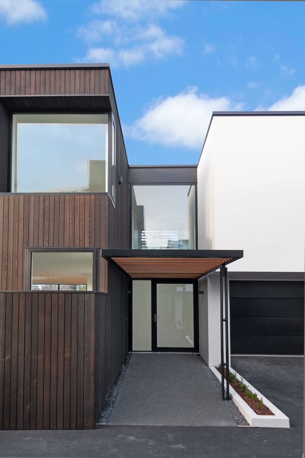 Exterior view of a contemporary multi-volume residence entry. The facade combines dark vertical timber cladding, smooth white rendered walls, black-framed glazing, and a projecting flat-roof entry canopy with a timber soffit. A paved approach leads to a glazed front door, with an attached dark garage visible to the right. The composition is clean and geometric under a bright blue sky.