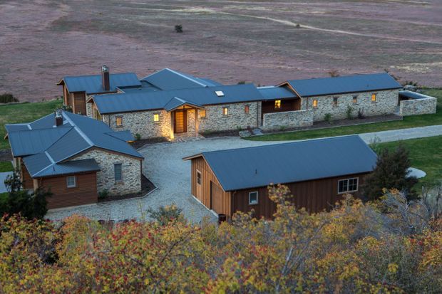 Aerial view of a low-rise rural mountain property comprising several connected and detached buildings. The structures feature dark blue-grey pitched metal roofs, natural stone walls, timber-clad sections, warm illuminated windows, and gravel driveways, set among autumn vegetation and open rolling terrain.