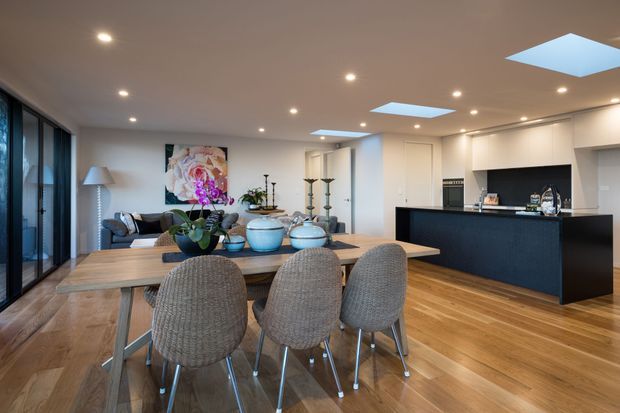 Open-plan contemporary kitchen, dining and living area with warm timber flooring, a large dining table, upholstered dining chairs, dark kitchen island cabinetry, white walls and ceiling, recessed lighting, and multiple skylights. Floor-to-ceiling glazing lines the left side, while a lounge area and decorative built-in shelving are visible beyond the dining zone.
