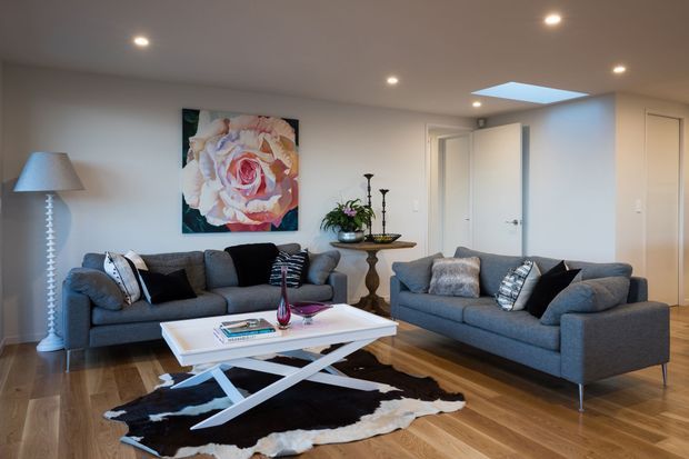 A bright, contemporary living room with two charcoal-grey sofas arranged around a white rectangular coffee table. The room features warm timber flooring, white walls and ceiling with recessed downlights, a large floral artwork, a sculptural floor lamp, decorative console table, patterned cushions, and a black-and-white cowhide rug. A roof skylight adds natural light, and doorways are visible at the rear.