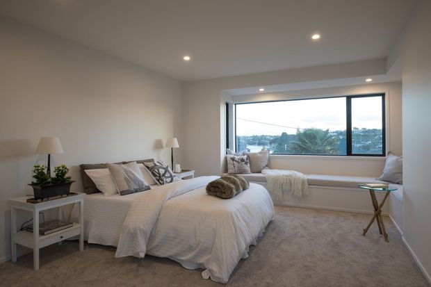 A bright, softly styled bedroom with two beds dressed in white bedding, neutral cushions, bedside tables and lamps, a broad built-in window seat, and a large horizontal window overlooking greenery and neighbouring rooftops. The room has recessed ceiling lights, pale walls, and plush beige carpeting, creating a calm and comfortable contemporary interior.