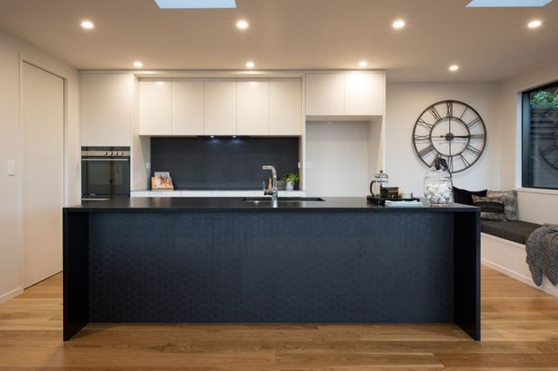 A contemporary open-plan kitchen dominated by a large dark island with an integrated sink and tap. White full-height cabinetry, a built-in oven, dark splashback, recessed ceiling lights, and skylights create a clean, modern fit-out. Timber flooring adds warmth, while a built-in window seat and oversized wall clock are visible in the adjoining living area.