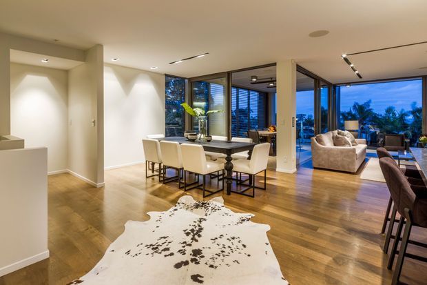 Open-plan contemporary interior combining a dining area and living room, with extensive floor-to-ceiling glazing opening toward a landscaped outdoor view. The space has warm timber flooring, white walls, recessed and track lighting, a dark dining table with upholstered cream chairs, and a pale sectional sofa. A cowhide rug adds a rustic accent to the otherwise refined modern setting.