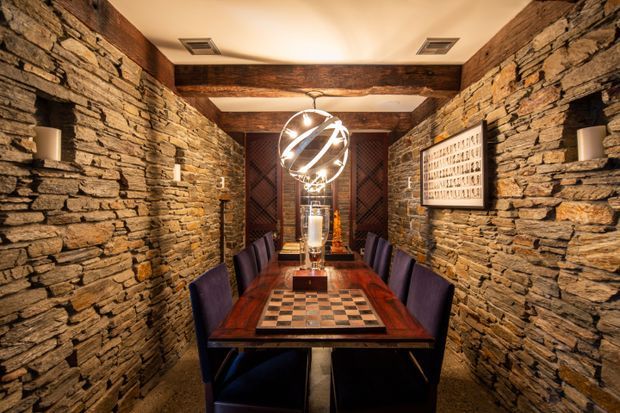 A narrow, dramatic dining room with full-height rough stacked-stone walls, exposed dark timber beams, and a long dark timber dining table surrounded by upholstered purple dining chairs. A large sculptural spherical pendant light hangs centrally above the table, with warm concealed ceiling lighting, decorative artwork, and a patterned tabletop creating a luxurious rustic atmosphere.