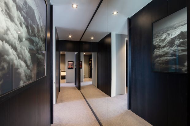 A contemporary interior hallway or entry corridor with dark timber wall panelling, full-height mirrored panels, recessed ceiling spotlights, and beige carpet. Large monochrome landscape artworks add visual interest, while a doorway at the rear leads toward another room.