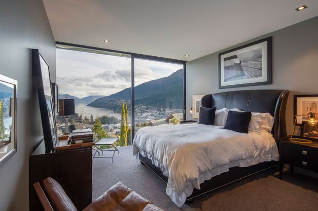 A refined contemporary bedroom with a large upholstered bed, dark bedside tables, soft neutral bedding, a leather lounge chair, and a wall-mounted television. Floor-to-ceiling corner glazing provides expansive views over a mountain-framed waterfront town. The room has recessed ceiling lighting, dark feature walls, and a calm, luxurious atmosphere.