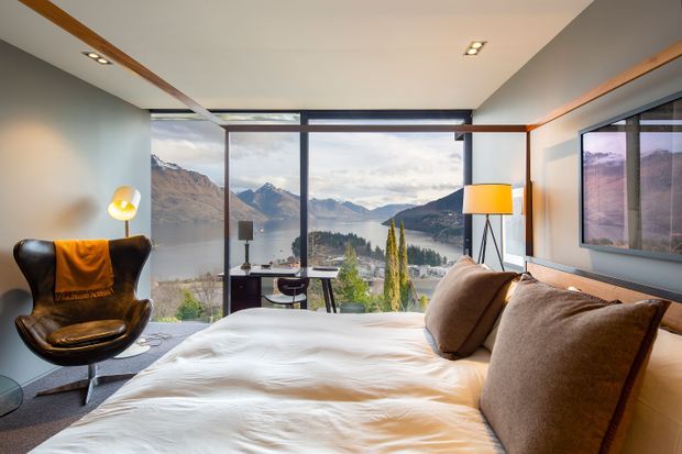A stylish bedroom with a large bed facing floor-to-ceiling glazing and a spectacular mountain-and-lake view. The room features warm timber ceiling accents, neutral walls, dark-framed windows, a leather lounge chair, bedside workspace, contemporary lighting, and a large framed landscape image on the wall.