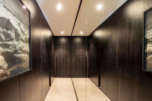 A narrow, luxurious lift or hallway lined with dark vertical timber wall panelling and large framed monochrome artwork. A mirrored rear wall visually extends the space, while a pale stone or terrazzo floor and recessed ceiling lighting provide contrast.