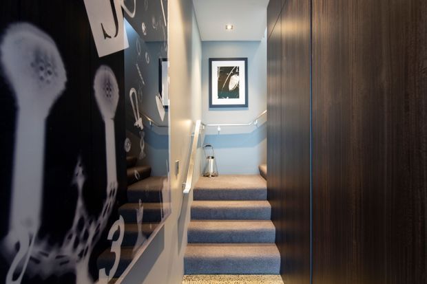 A narrow interior stairway with blue carpeted steps, pale blue walls, white trim and handrails, and dark wood wall panelling. Large monochrome graphic artwork and framed art add a bold decorative element, while a small landing and upper hallway are visible beyond the stairs.