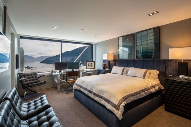 A sophisticated modern bedroom with a large upholstered bed, dark leather tufted lounge chairs, integrated bedside furniture, and a compact work area by expansive floor-to-ceiling glazing. The room overlooks a mountainous waterfront landscape and uses a restrained palette of charcoal, black, white, grey, and warm wood tones, with sleek dark cabinetry and contemporary recessed lighting.