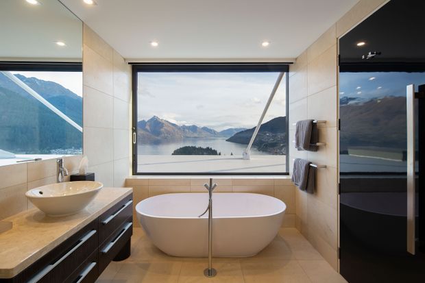 A contemporary bathroom with a freestanding oval bathtub positioned in front of expansive picture windows overlooking a dramatic alpine lake and mountains. The room features beige tiled walls and floor, a timber-toned stone-look vanity benchtop with dark built-in cabinetry, a vessel basin, chrome tapware, wall-mounted towel rails, recessed ceiling lights, and a glossy dark-framed door or partition.