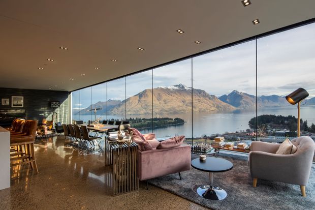 A spacious, luxurious living room with floor-to-ceiling glazing overlooking a lake and dramatic mountainous landscape. The room features polished speckled stone or terrazzo flooring, dark feature wall panelling, a dining area, contemporary sofas and armchairs, a sculptural pendant or floor lamp, and recessed ceiling lighting.