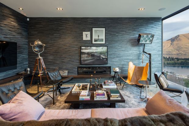 A stylish living room with dark stacked-stone feature walls, a low contemporary media console, leather lounge chairs, a large sectional sofa, and a central coffee table arranged with books and decor. Recessed ceiling lights provide warm illumination, while full-height glazing at right offers a scenic mountain and lake view.
