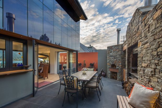 A sophisticated enclosed courtyard terrace beside a contemporary glass-walled building and substantial dry-stone masonry walls. The paved outdoor dining area has a rectangular table, ornate metal chairs, built-in bench seating with cushions, a barbecue or fireplace zone, and a warm red rendered feature wall. Large glazed openings connect the terrace to the interior, while exposed chimneys and roof forms add architectural character.