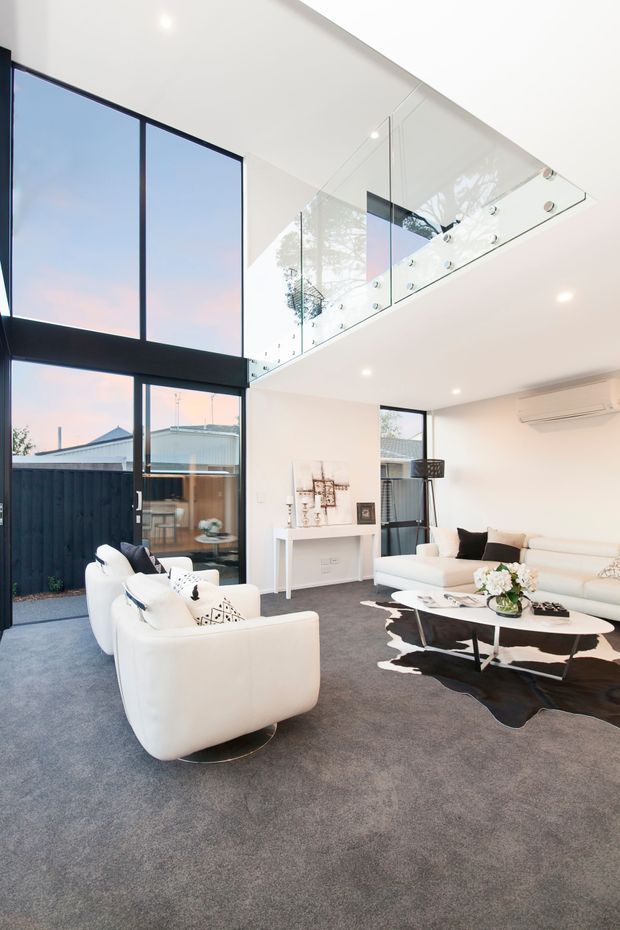 Bright contemporary living room with a double-height glazed wall, large black-framed windows and doors, a clear glass balustrade overlooking the space, white sectional sofa and lounge chairs, dark oval coffee tables, and a grey carpet. A fireplace wall and outdoor courtyard are visible beyond the glazing.