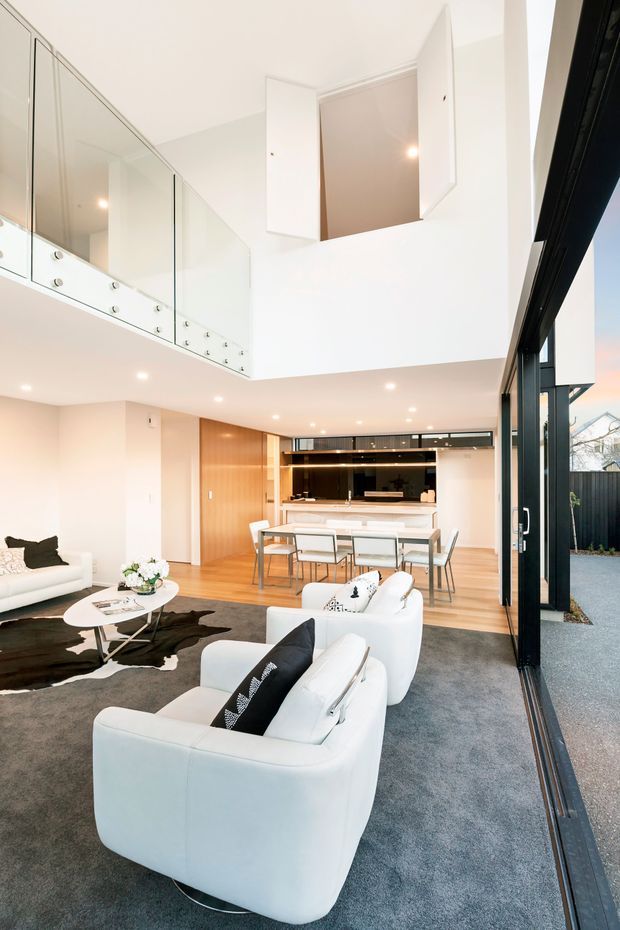 Bright contemporary open-plan living, dining and kitchen interior with a double-height void and mezzanine-style upper landing. White curved sofas sit on a grey carpet beside a cowhide rug and round coffee table, while the dining area and streamlined kitchen occupy the rear. Full-height black-framed glazing opens to an exterior paved area, and glossy white cabinetry with glass balustrading lines the upper level.