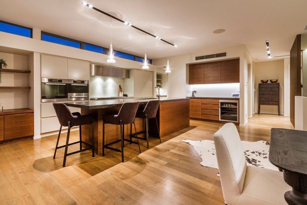 Open-plan contemporary kitchen and dining area with warm timber flooring, light neutral cabinetry, a large island with bar seating, integrated stainless-steel appliances, pendant lights, and a timber dining table in the foreground. Blue clerestory windows add a vivid accent, while a built-in wine refrigerator and timber joinery are visible beyond the island.