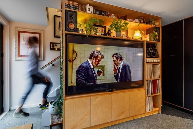 A warm, built-in timber entertainment wall dominates the room, with a large television set into open shelving and lower cabinetry. The shelving displays books, records, speakers, framed objects, and abundant potted greenery. A dark painted wall and carpeted floor create contrast, while a blurred person moves through the adjacent hallway or entry area.