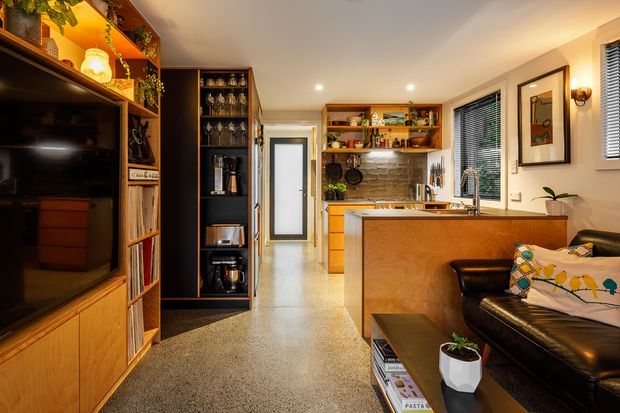 Warm, compact open-plan living and kitchen interior with a timber media wall and shelving on the left, a dark leather sofa in the foreground, and a timber kitchen island and cabinetry at right. The kitchen includes open display shelving, stainless fixtures, a tiled backsplash, and a window with blinds. Polished concrete or terrazzo-like flooring, recessed lighting, plants, books, and decorative objects create a refined, lived-in atmosphere.