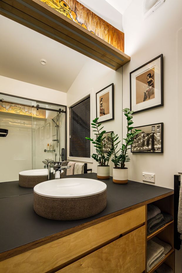 A warm, contemporary bathroom vanity with a large round vessel basin on a dark benchtop, timber drawer cabinetry, open towel storage, and a matching basin reflected in the mirror. A glass-enclosed shower, black venetian blind, framed artwork, indoor plants, and timber detailing complete the space.