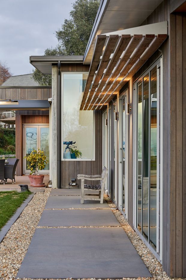 A modern residential exterior walkway beside a timber-clad building. The covered passage features full-height glazed doors and windows, exposed timber pergola rafters, vertical wood cladding, concrete stepping slabs, and gravel landscaping. A small courtyard and patio area are visible beyond.