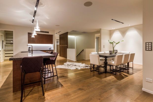 Open-plan contemporary kitchen and dining area with warm timber flooring, a large dark island with seating, integrated cabinetry, pendant lighting, and a six-seat dining table. A stairwell and built-in storage are visible in the background. The space has a warm, refined, softly lit atmosphere.