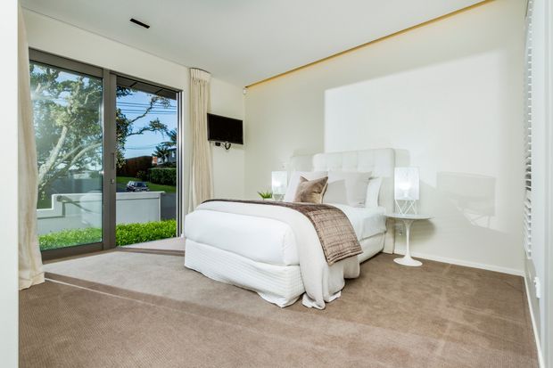 Bright, refined bedroom with a white upholstered bed, layered white bedding, and a taupe-brown throw. The room has wall-to-wall beige carpet, white walls and ceiling, a slim brass-toned ceiling trim, bedside tables with lamps, and a wall-mounted television. Full-height sliding glass doors open to a small landscaped outdoor area, bringing in abundant natural light.