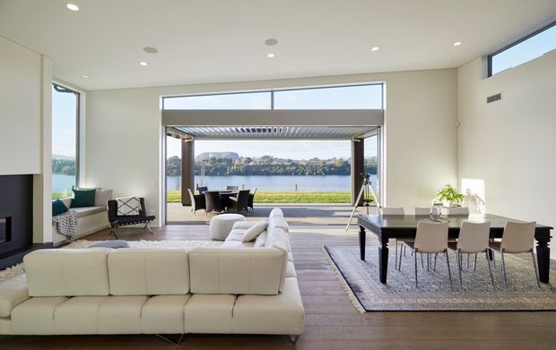 Bright contemporary open-plan living and dining room with a large cream sectional sofa, dark dining table and upholstered chairs, timber flooring, and extensive floor-to-ceiling glazing opening onto a covered terrace. The room overlooks a calm body of water and a landscaped shoreline, with minimalist recessed lighting and neutral finishes.