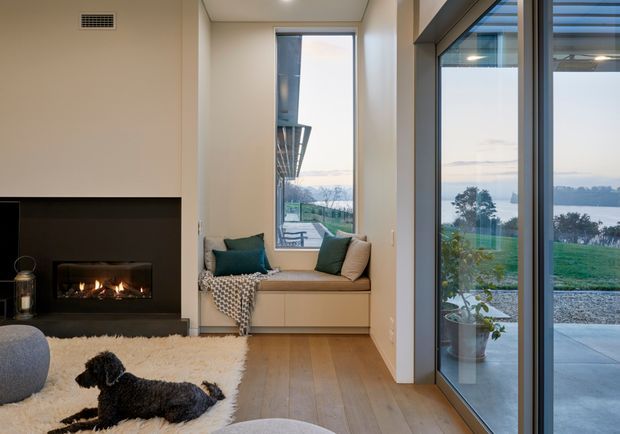 A warm, contemporary living room with a built-in black fireplace, timber flooring, and a window seat overlooking a landscaped waterfront or lakeside setting. Full-height glazed doors and windows bring in abundant natural light, while neutral walls, teal cushions, and a textured throw add contrast. A dog rests on a pale rug in the foreground.