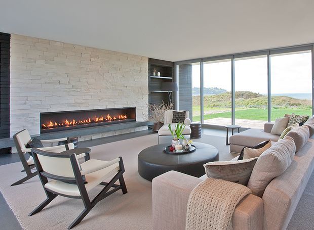 A spacious contemporary living room with a long linear built-in fireplace set into a pale textured stone feature wall. Floor-to-ceiling glazing opens to a coastal landscape, while a large sectional sofa, sculptural round coffee table, armchairs, and built-in shelving create a refined, comfortable seating area. The palette is soft beige, cream, grey, and natural timber tones with abundant daylight.