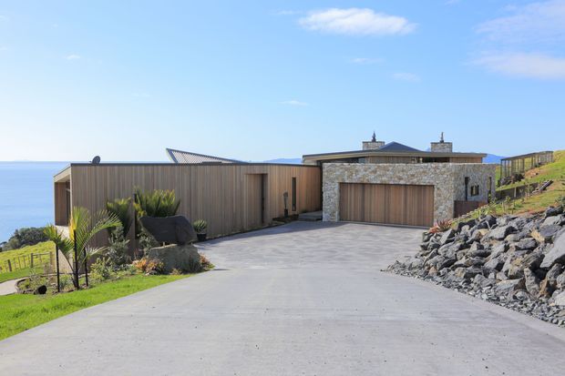 A contemporary coastal residence viewed from its broad concrete driveway. The low, angular house combines vertical timber-look cladding with a substantial rough-stone garage facade and a timber garage door. Landscaping, exposed rock retaining areas, fencing and ocean views reinforce the dramatic sloping coastal setting.