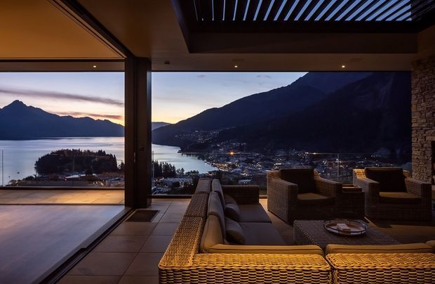 A luxurious covered outdoor lounge or terrace at dusk, furnished with woven wicker seating and cushions, overlooking a lake, illuminated settlement, and dramatic mountainous landscape. Large open glazing connects the lounge to an adjoining interior, while a dark slatted overhead canopy and stacked-stone wall frame the view.