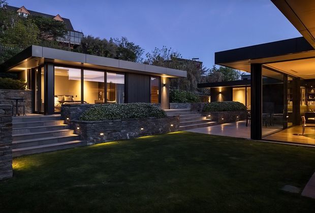 Twilight view of a modern multi-level residence arranged around a landscaped lawn and terrace. The low, flat-roofed pavilion has dark cladding, extensive floor-to-ceiling glazing, warm concealed lighting, broad stone steps, raised planter walls, clipped hedges, and a glazed entertaining pavilion on the right with visible bar joinery. A larger house is partially visible uphill behind the main structure.
