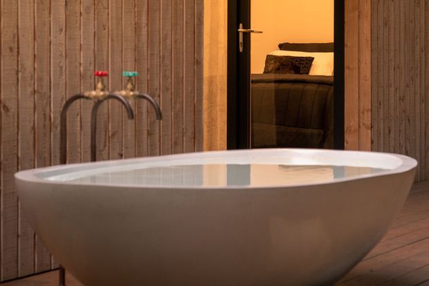 A freestanding oval bathtub dominates an intimate bathroom setting, positioned against vertical timber wall cladding and a timber plank floor. Two curved wall-mounted taps are visible behind the tub, while an open dark-framed doorway reveals part of a warmly lit bedroom with a bed beyond.
