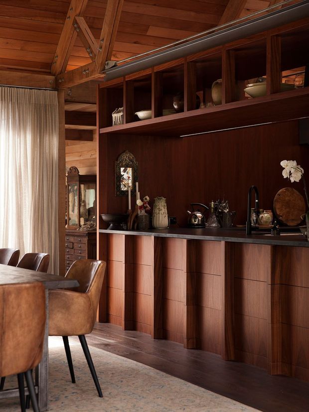 A richly timber-lined dining area with a built-in dark wood drinks and serving cabinet, open display shelving, sink, and dark benchtop. Leather dining chairs and part of a dining table sit in the foreground. The room features exposed timber ceiling beams and wall cladding, warm textured finishes, and a tall sheer-curtained window.