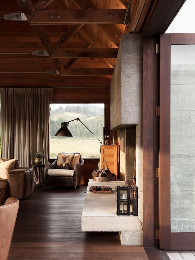 Warm rustic living room with dark timber flooring, exposed timber trusses and ceiling, heavy drapery, leather and upholstered lounge seating, and a substantial concrete or masonry fireplace with a projecting hearth. Large glazing opens views toward a rural landscape, while a dark timber-framed door or window dominates the right side.