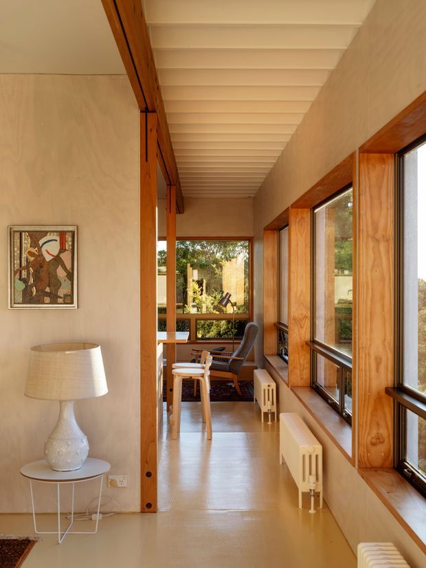 A warm, narrow glazed corridor or landing with extensive timber-framed windows on one side, a pale smooth floor and walls, and exposed white-painted ceiling rafters. The space contains a small dining or work table with chairs at the far end, a console table with lamp and artwork, and radiators beneath the windows. Mature greenery is visible outside through the glazing.