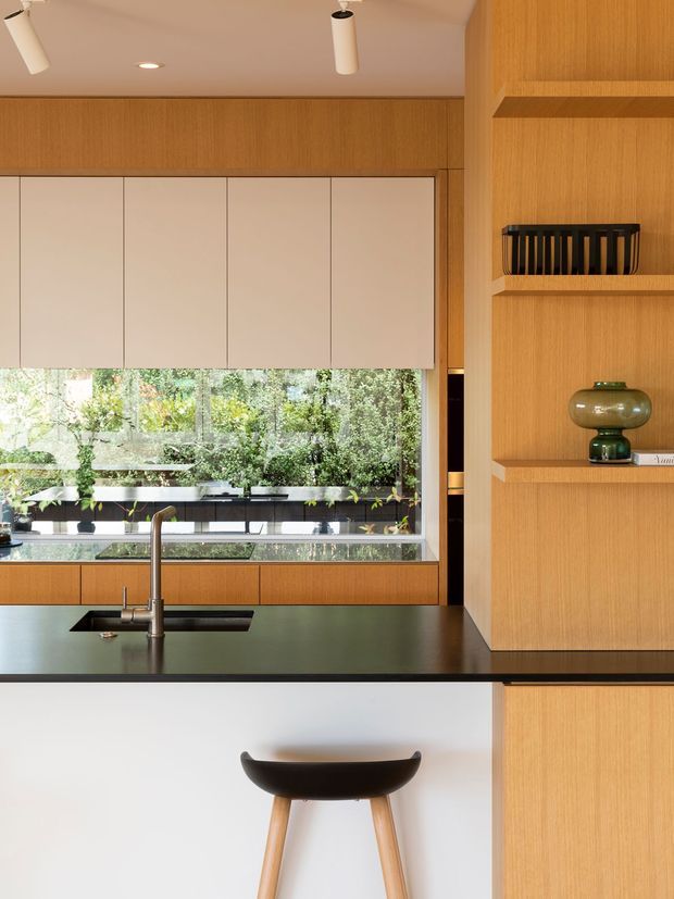 A refined contemporary kitchen with warm timber cabinetry and integrated open shelving, matte light upper cabinets, and a dark stone island benchtop with an undermount sink and tall brushed-metal tap. A large window provides views toward lush greenery, while a sculptural timber-legged bar stool sits at the island. Recessed and cylindrical pendant lighting complete the clean, understated composition.
