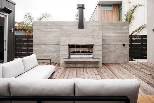 Contemporary outdoor entertaining area with a built-in linear gas fireplace set into a horizontal grey tiled or concrete-look feature wall, with a black flue above. A timber deck, white sectional sofa, dark fencing, and surrounding modern residences create a refined courtyard-like setting.