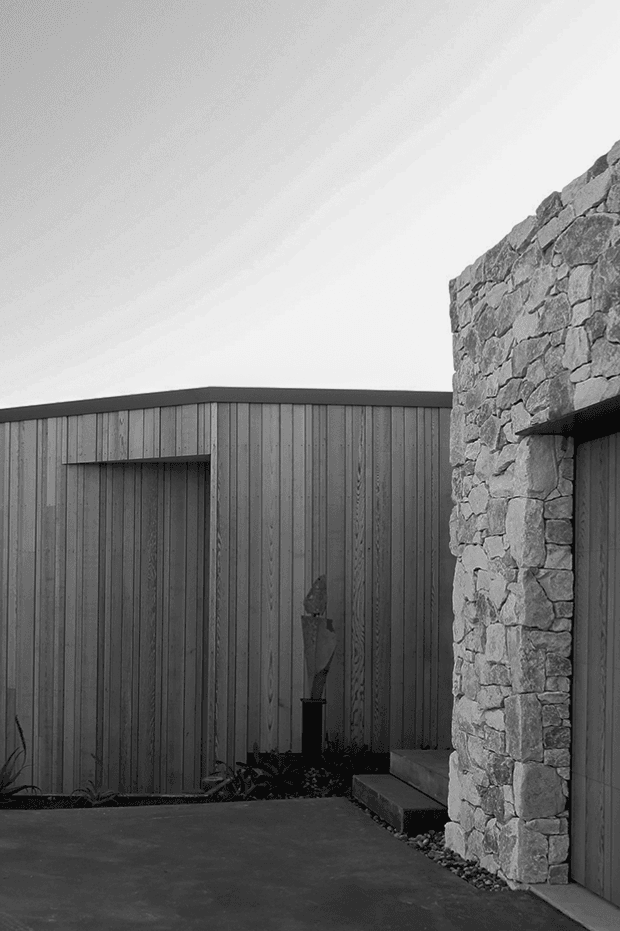 Black-and-white architectural exterior photograph showing a minimalist courtyard or entry approach. A dark vertically clad timber building with a low flat roof meets a prominent rough natural-stone wall, with a narrow paved ground plane and simple planted edge. The composition emphasises contrasting textures between weathered stone and vertical timber cladding.