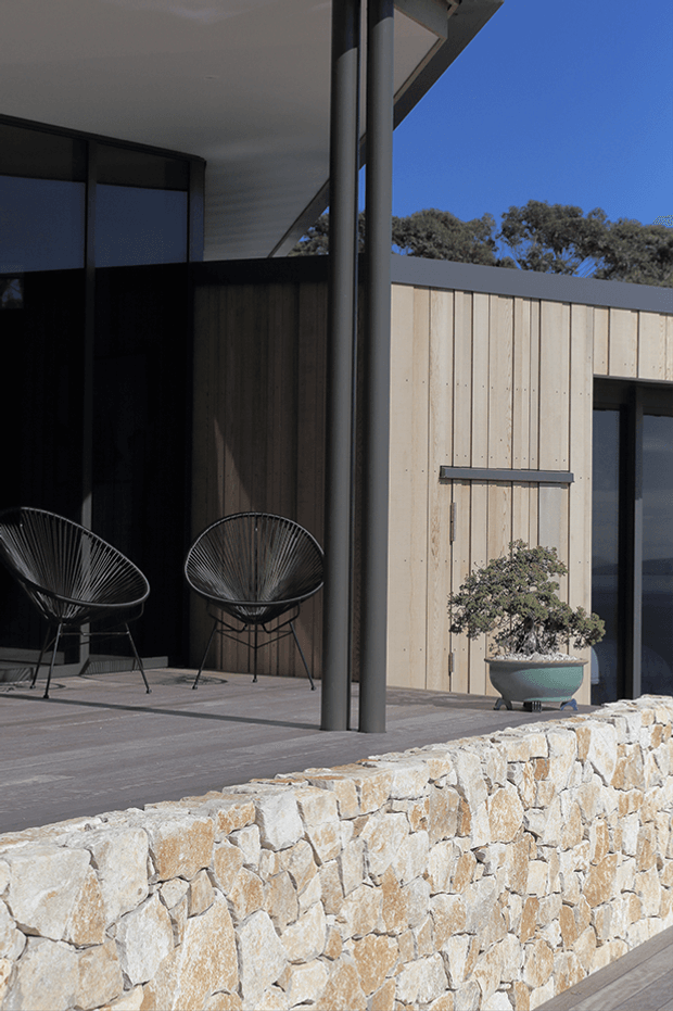 Exterior view of a contemporary single-level building with pale vertical timber cladding, dark metal-framed glazing, a broad roof overhang, and slender dark structural columns. A paved terrace with two black wire lounge chairs overlooks a rough natural-stone retaining wall, with a sculpted potted tree near the entrance. The composition highlights the facade, terrace, and material contrast under clear daylight.