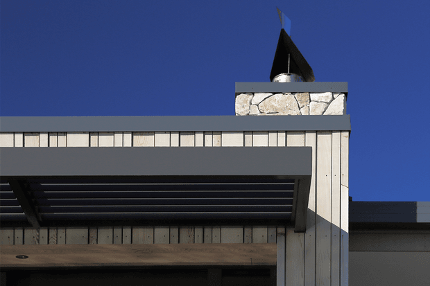 Close-up exterior view of a contemporary building facade under a deep blue sky. The composition shows pale vertical cladding, dark grey horizontal bands, a projecting flat-roofed canopy with dark slatted underside, and a stone-clad chimney or tower topped by a dark angular metal cap. The image focuses on architectural materials, roofline, and strong geometric detailing.