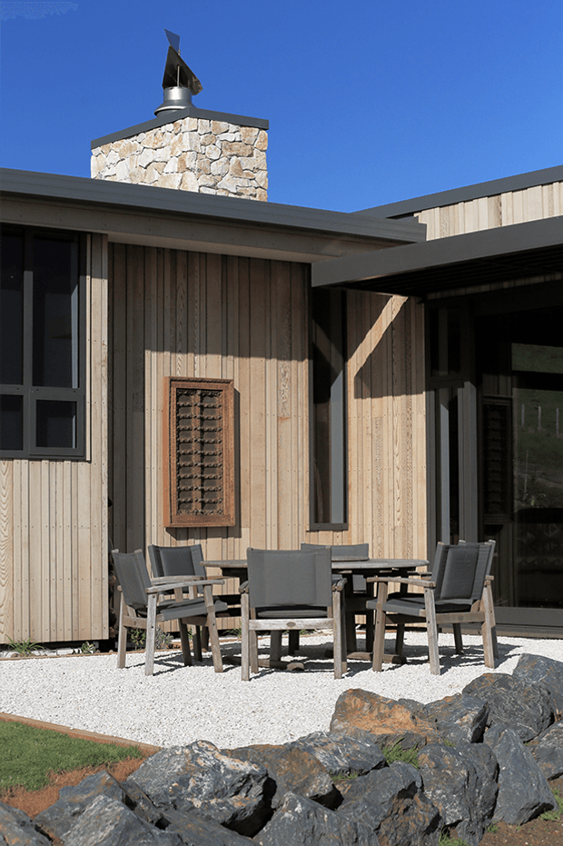 Exterior view of a contemporary single-storey building with pale vertical timber cladding, dark metal roof edges and window frames, and a prominent rough-stone chimney with metal cap. A covered outdoor seating area with a table and chairs sits on a white gravel terrace bordered by dark rocks. Strong sunlight creates crisp shadows against a clear blue sky.