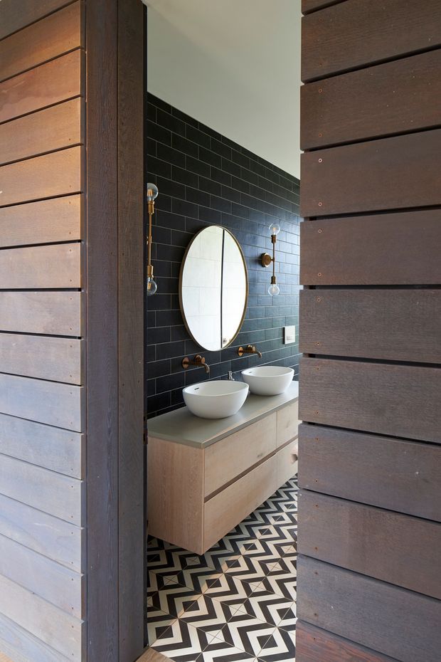 A compact, contemporary bathroom viewed through a dark timber doorway. The room features a double vanity with two white vessel basins, a pale timber floating cabinet, matte black subway-tile walls, brass wall-mounted taps and sconces, a round mirror, and a bold black-and-white geometric patterned floor. Dark horizontal timber cladding frames the opening.