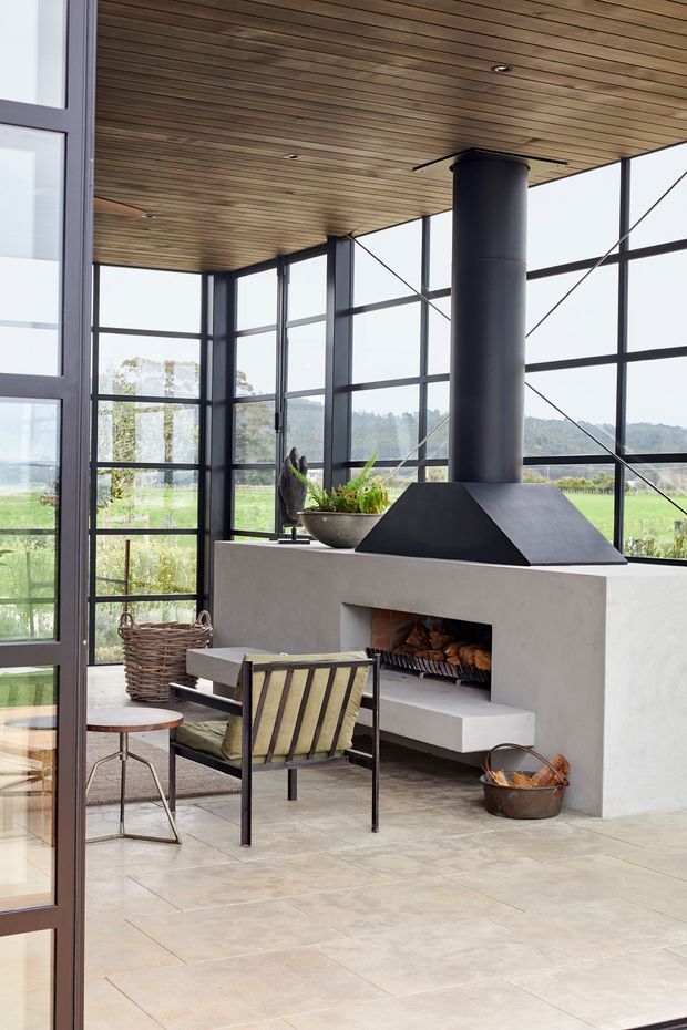 A bright, glazed indoor-outdoor living space with floor-to-ceiling black-framed windows, a warm timber-lined ceiling, pale tiled flooring, and a substantial built-in concrete fireplace with a black metal hood and flue. Lounge chairs, a small side table, firewood, and a woven basket furnish the room, while a green rural landscape is visible through the glazing.