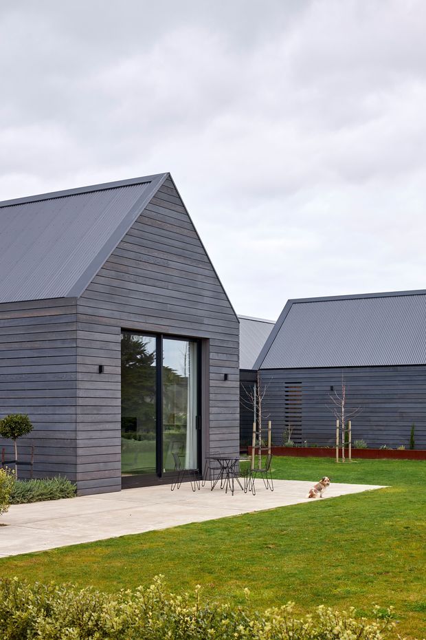 Exterior view of a contemporary cluster of gabled buildings with dark horizontal timber cladding and corrugated metal roofs. Large glazed doors open onto a pale concrete patio, with a neatly landscaped lawn and young trees in the foreground. The composition emphasizes the pitched roof forms, restrained dark palette, and connection between the buildings and outdoor space.